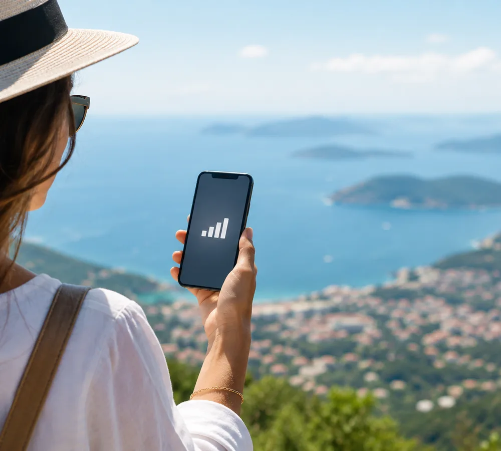 Voyageuse au chapeau tenant son iPhone face à une baie et ses îles, réseau au maximum sur l’écran